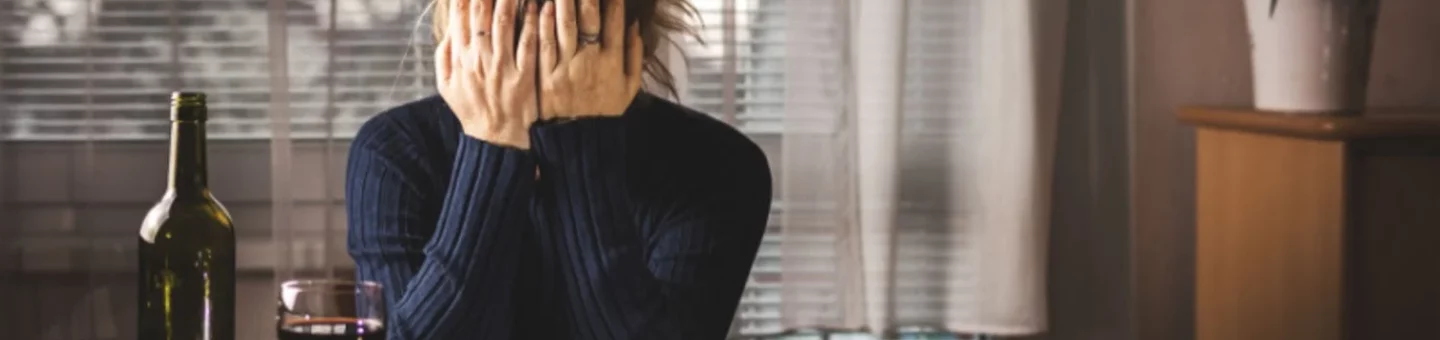 Distressed woman holds head in hands with a bottle of wine and pills in front of her