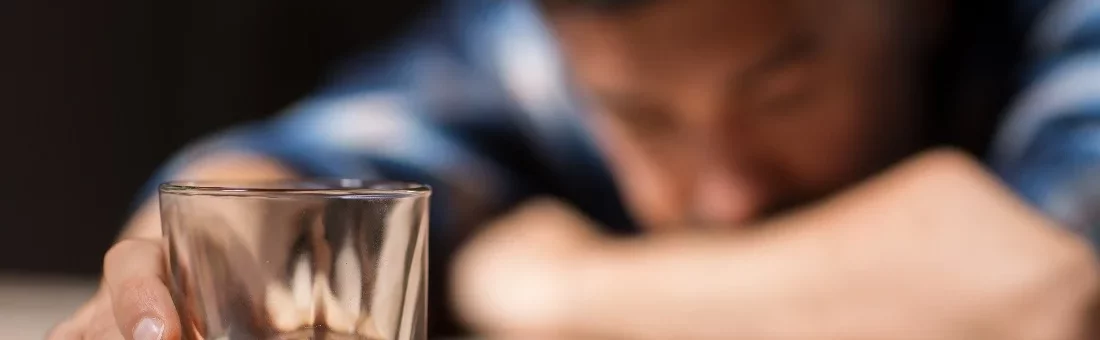 Man with his head on the table rests his hand on a glass of alcohol