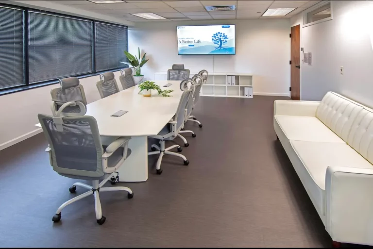 Meeting room with office chairs set around a large round table, a television mounted on the wall, and white leather couch at Legacy Healing Center’s Parsippany drug and alcohol rehab