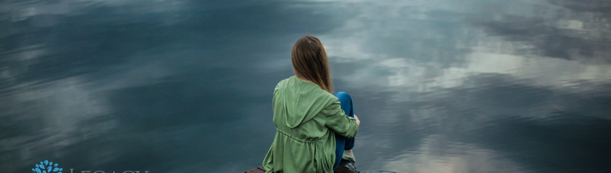 A woman sits on a dock next to a lake - stares off into the distance.